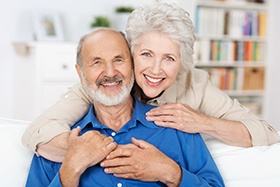 An older man and woman seated on a couch and smiling after receiving dental implants