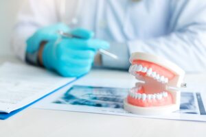 A desk with a set of X-rays, paperwork, and model dentures in front of a dentist with blue gloves who is blurry in the background